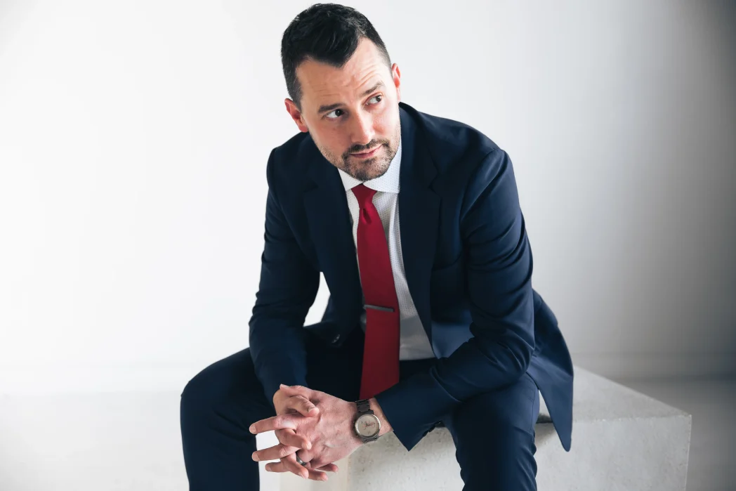 Josh Fletcher mentalist seated in a navy suit and red tie with hands clasped