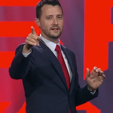 Josh Fletcher performing on stage in a suit with a red tie, pointing toward the audience